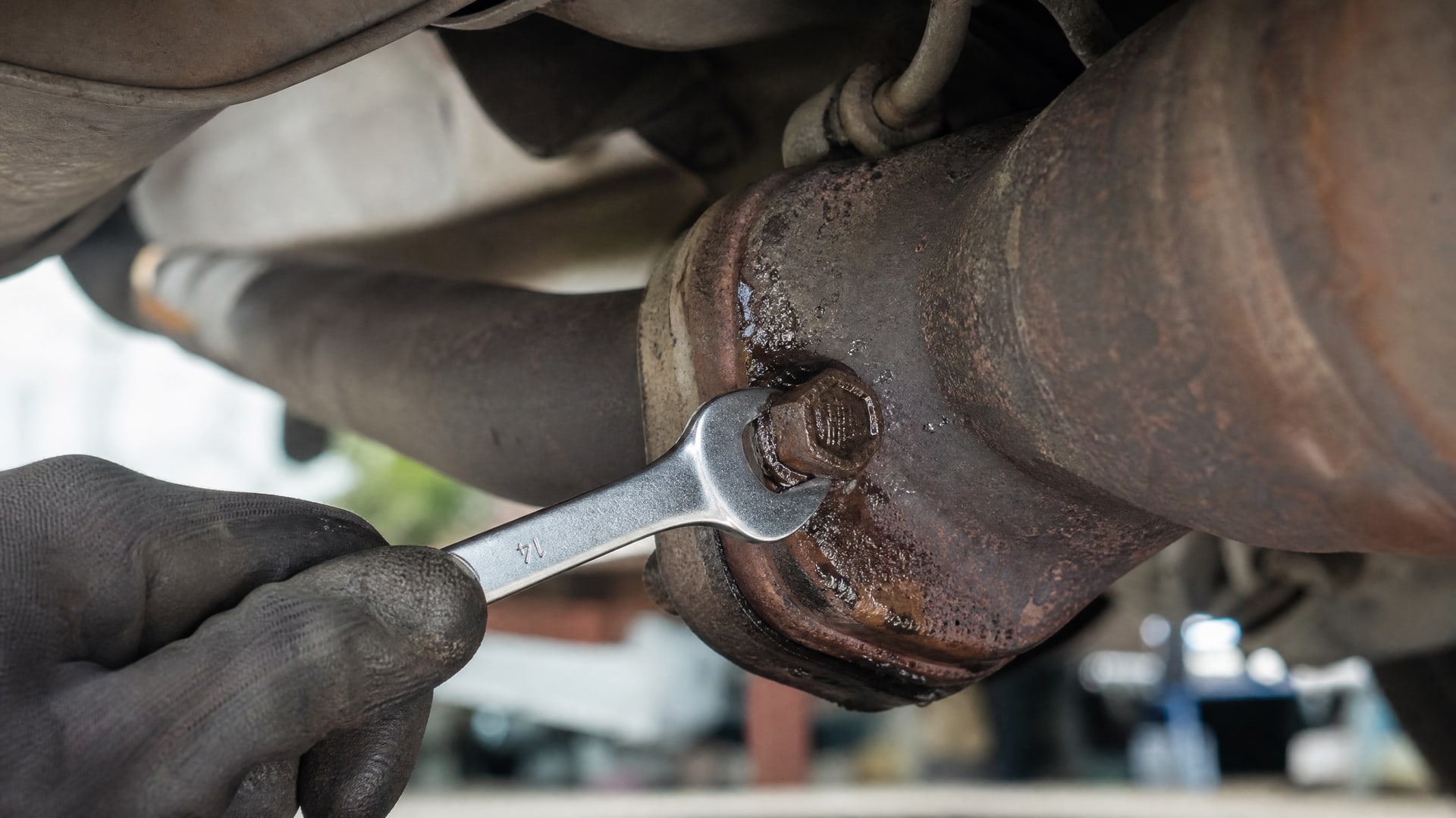 Рука механика медленно откручивает ржавый болт глушителя, смазанный WD-40.