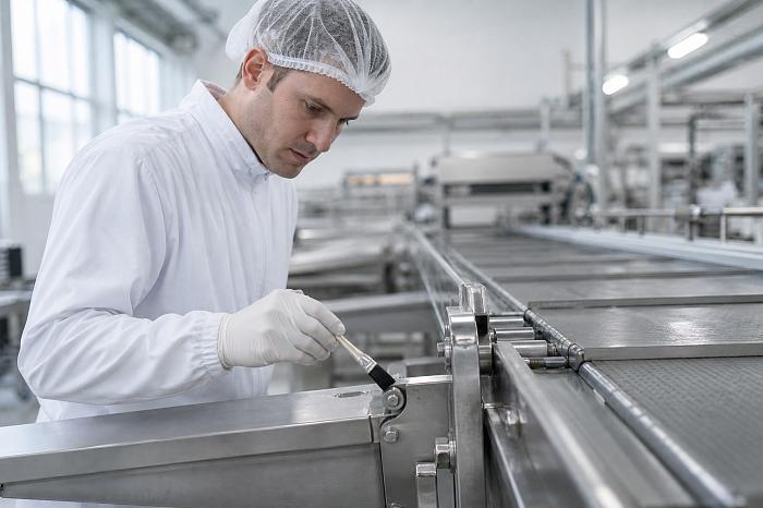 Техник в белом комбинезоне аккуратно наносит смазку пищевую nsf h1 на шарнир конвейерной ленты.
